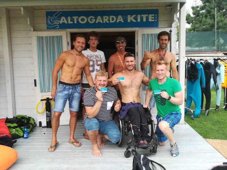 Ein Rollstuhlfahrer und zwei weitere Kiteschüler zeigen die frisch erworbene Mitgliedskarte. Auch Gründungsmitglieder des Vereins lächeln in die Kamera. Im Hintergrund das Gartenhäuschen des Vereins mit dem Schriftzug 'Altogarda Kite'.