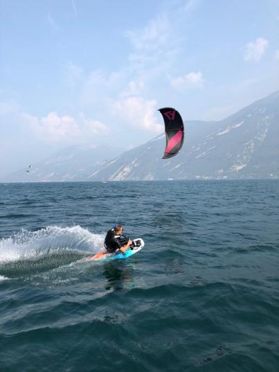 Ein Sitkiteboarder auf dem Wasser. Hinter dem Board sieht man den Spray des Wassers. Im Hintergrund ist der Kite zu sehen.