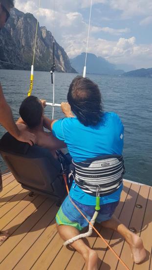 Ein Kiteschüler sitzt sicher in einem Autositz auf dem Boot und lernt die Kite-Steuerung. Der Kitelehrer kniet daneben und unterstützt den Schützling.