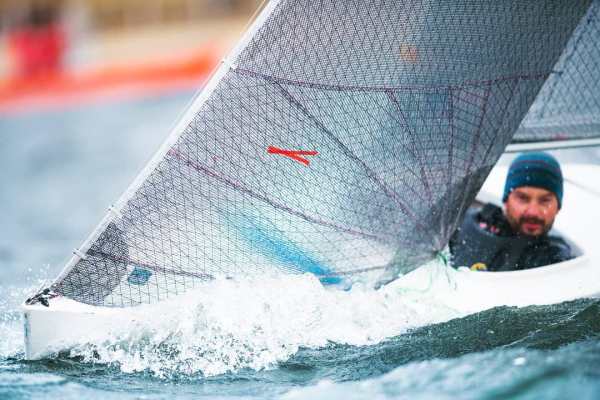 Nahaufnahme Segelboot in voller Fahrt. Das Segel ist vom Wind gebläht. Recht im Hintergrund sieht man den Segler etwas unscharf mit blauer Mütze.