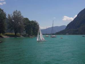 See bei Sonnenschein. Im Zentrum ein 2.4mR Boot. Im Hintergrund sieht man ein paar geankerte Boote sowie das Ufer mit Bäumen und Gebirge in der Ferne.