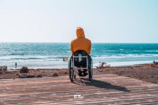 Wheelchair user with a yellow hoody on a wooden plattform on the beach. The photo is taken from behind. The hood covering the head. In the background is the sea with one windsurfer on the shore.