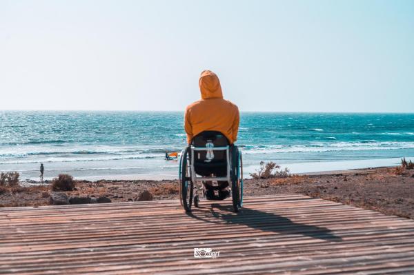 Wheelchair user with a yellow hoody on a wooden plattform on the beach. The photo is taken from behind. The hood covering the head. In the background is the sea with one windsurfer on the shore.