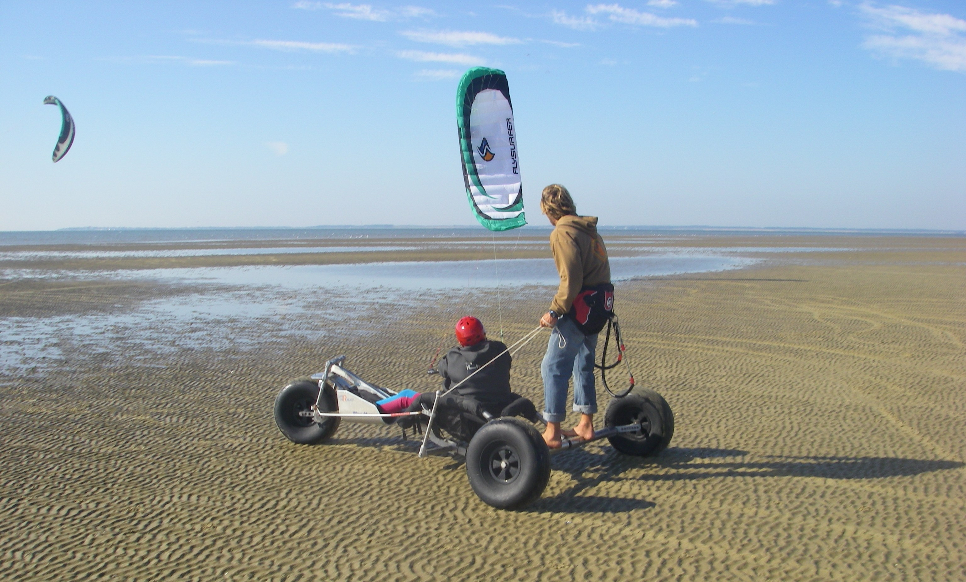 A person with red helmet sits in an adapted kitebuggy. Behind him on the axle of the buggy stands a man to help with the steering.