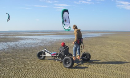 A person with red helmet sits in an adapted kitebuggy. Behind him on the axle of the buggy stands a man to help with the steering.