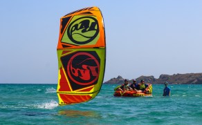 In front a red and yellow kite. In the background there is an inflateable couch in the sea - 3 people sitting on top. A 4th person stands next to them in the shallow water.