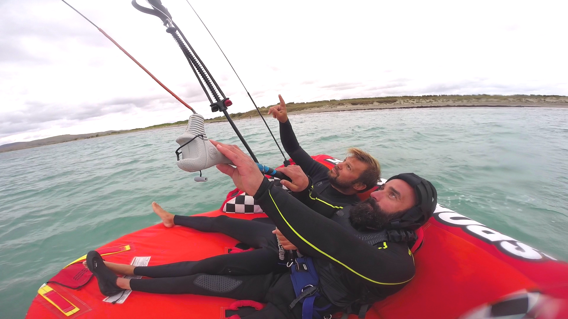 Two men on an inflateable sofa on the water. The man in front with beard and helmet is holding a kite-bar. The man in the background is pointing to the kite.
