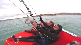Two men on an inflateable sofa on the water. The man in front with beard and helmet is holding a kite-bar. The man in the background is pointing to the kite.