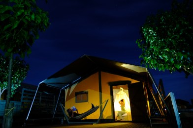 An illuminated tent with the door open. Inside a woman sits on a bed. Outside on the dark porch a man is lying in a hammock in the dark of the night.