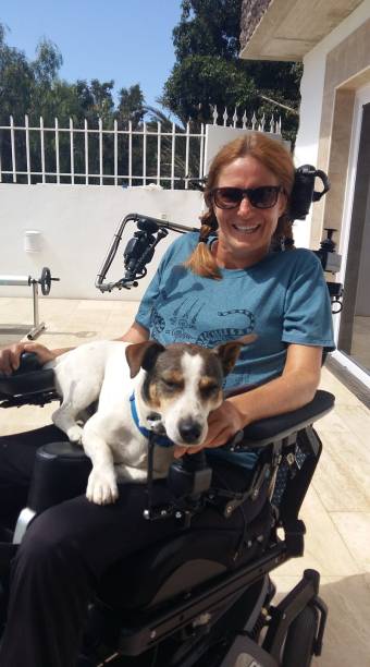 Woman in an electric wheelchair wearing sunglasses. A small dog sits on her lap.