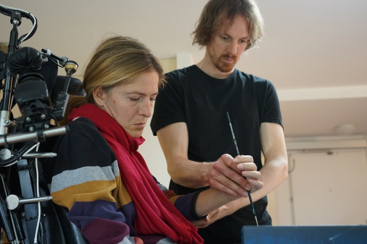 Woman in an electric wheelchair. To her left stand a man with longer hair helping her to hold a brush for painting.